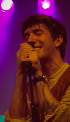 a man singing into a microphone in front of purple and pink lights on stage at a concert