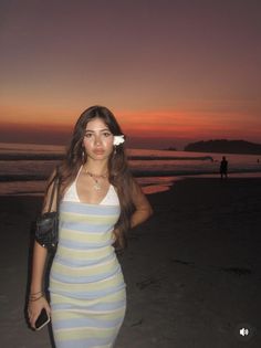 a woman standing on top of a beach next to the ocean at sunset with her hands in her pockets