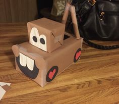 a cardboard box shaped like a truck with eyes and lips on it sitting on a wooden floor next to a handbag