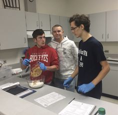 three young men standing around a table with food in a mixing bowl and gloves on