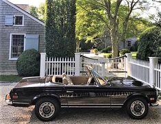 a black convertible car parked in front of a white picket fence with a dog sitting in the driver's seat