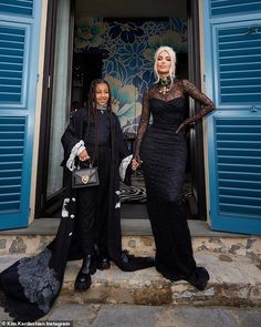 two women standing in front of a door with blue shutters on the outside and one wearing a black dress