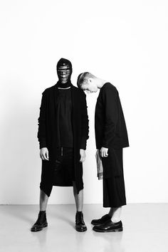 two men standing next to each other in front of a white wall wearing black clothing