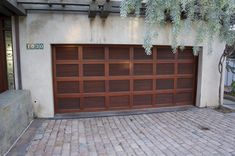 a brown garage door sitting on top of a brick floor next to a tree and bushes