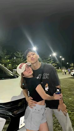 a man and woman standing in front of a car at night with their arms around each other