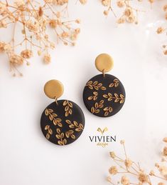 two black and gold earrings with leaves on them next to some dried flowers in front of a white background