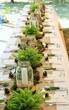 a long table is set with pine cones, ferns and candles for an outdoor dinner