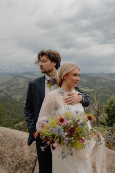 a man and woman standing next to each other on top of a hill with mountains in the background