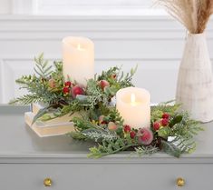 two white candles sitting on top of a table next to a vase filled with berries and greenery