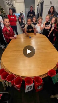 a group of children standing around a wooden table with cups on it and people in the background