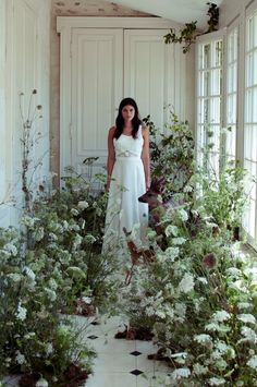 a woman in a white dress standing next to some plants and flowers on the floor