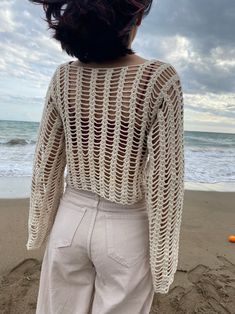 a woman standing on top of a sandy beach next to the ocean wearing a white crocheted sweater