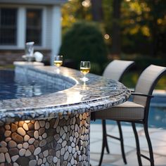 two wine glasses sitting on top of a table next to a swimming pool with chairs around it