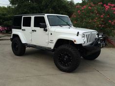 a white jeep parked in a driveway next to some bushes and trees with pink flowers behind it