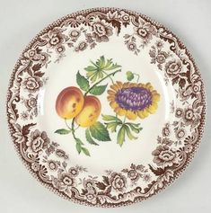 an ornately decorated plate with fruit and flowers on the rim, against a white background