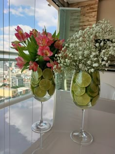 two wine glasses with flowers and limes in them on a table next to a window