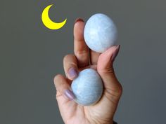 a hand holding two blue and white balls in front of a yellow crescent moon on a gray background