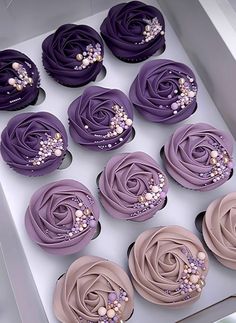 twelve cupcakes with purple frosting and pearls on the top are arranged in a box