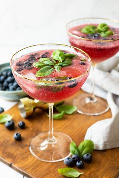 two glasses filled with blueberries and basil on top of a cutting board next to another glass