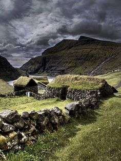 an old stone house with grass roofs on the side of a hill near water and mountains