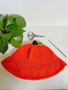 an orange crocheted hat sits next to a green plant and some scissors on a white surface