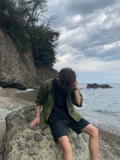 a man sitting on top of a rock next to the ocean while talking on a cell phone