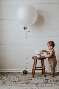 a baby standing in front of a table with a cake on it and a white balloon