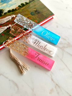 three personalized bookmarks with tassels are on a marble countertop next to an open book