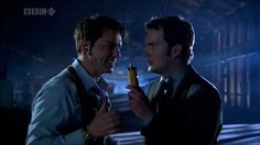 two men standing next to each other in a dark room with one holding a lighter