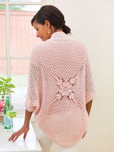 a woman standing in front of a window wearing a pink crocheted sweater and white pants