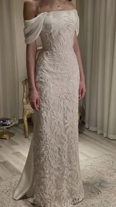 a woman standing in front of a window wearing a wedding dress with off the shoulder sleeves