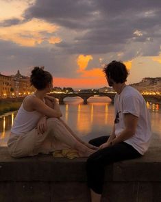 two people are sitting on a ledge near the water at sunset or dawn, with buildings in the background