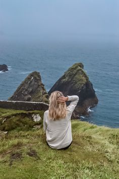 a woman sitting on top of a grass covered hill next to the ocean with her hands behind her head