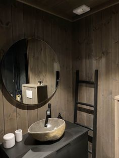 a bathroom sink sitting under a round mirror next to a wooden chair and wall mounted faucet