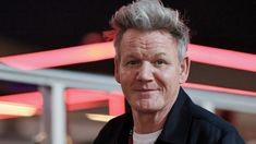 a man with grey hair standing in front of a neon sign and looking at the camera