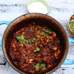 a pot filled with meat and beans next to a bowl of salad on top of a wooden table