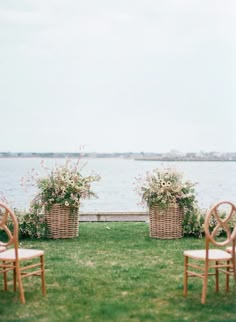 two wicker baskets with flowers are sitting on the grass near some chairs in front of water