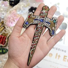 a hand holding a cross shaped brooch in it's left hand, with other jewelry on the table behind it