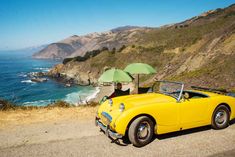 an old yellow sports car parked on the side of the road next to the ocean