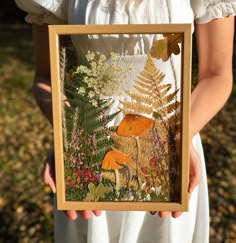 a woman holding up a framed painting in front of her face with leaves and flowers on it