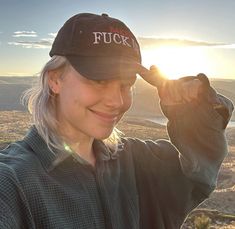 a woman with blonde hair wearing a black hat and smiling at the camera while standing on top of a hill