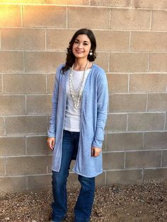 a woman standing in front of a brick wall wearing jeans and a long cardigan