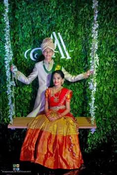 a man and woman sitting on a swing in front of a wall with greenery