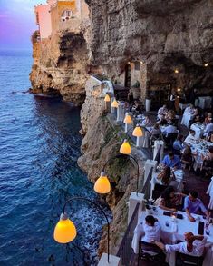 people are sitting at tables on the cliff by the ocean with lights in front of them