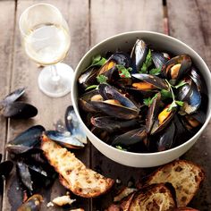 a bowl of mussels and bread on a wooden table next to a glass of wine