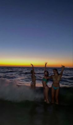 two women in bikinis standing on the beach with their arms up as the sun sets