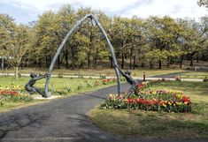 two statues in the middle of a park with flowers and trees around them, one is swinging on a pole