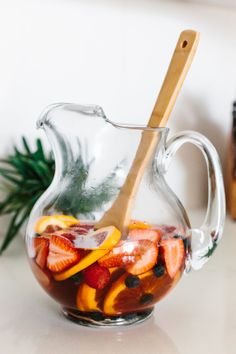 a glass pitcher filled with sliced strawberries and oranges next to a wooden spoon