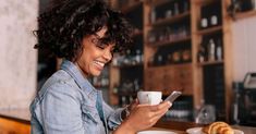 a woman sitting at a table using her cell phone and holding a cup of coffee