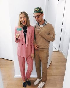 a man and woman standing next to each other in front of a white wall holding drinks